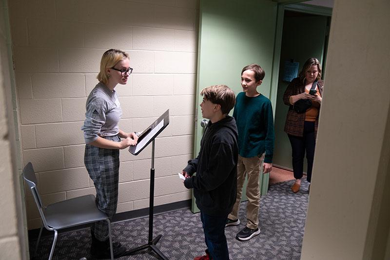 Student at a music stand listening to child talking holding a paper card. He stands next to another child. A woman looks at her phone in the doorway.